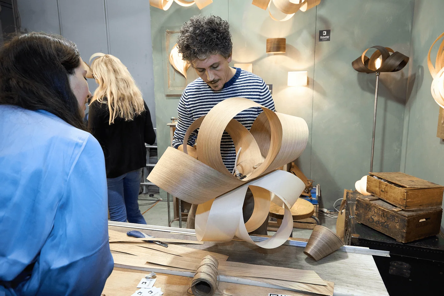 A craftsperson works on a wooden lamp in front of visitors.